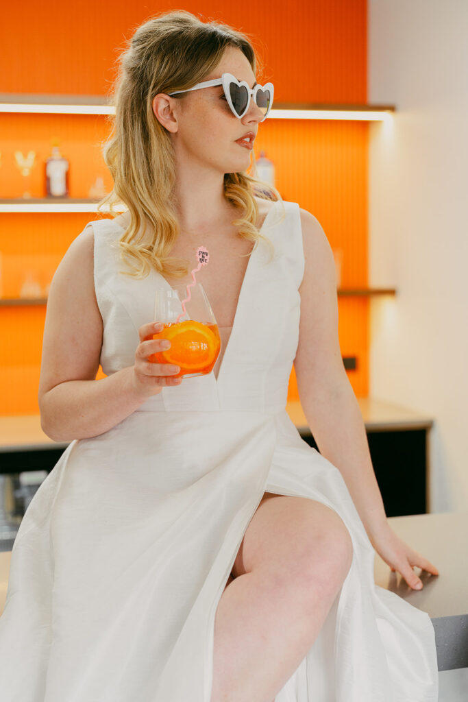 Beautiful bride sitting on the bar with heart sunglasses on and a glass of aperol spritz in her hand. Pink swizzle stick reads "crazy in love"