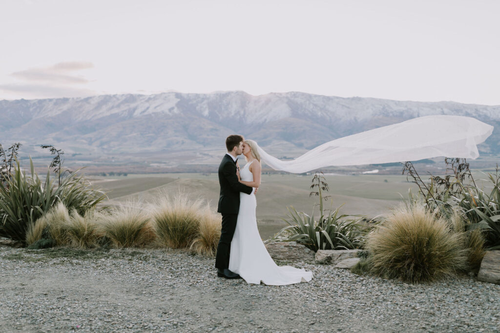 Bride and groom kissing with Central Otago mountains in the background and veil flying in the wind