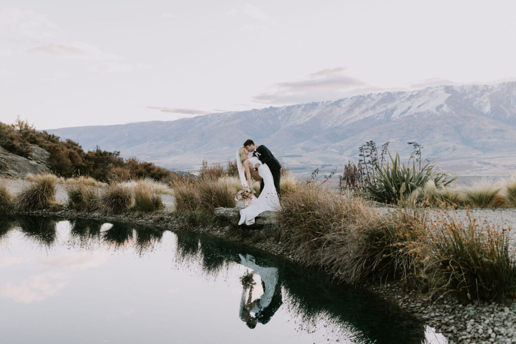 Bride and groom in a dip kiss with The Canyon pond in the foreground and Central Otago mountains in the background