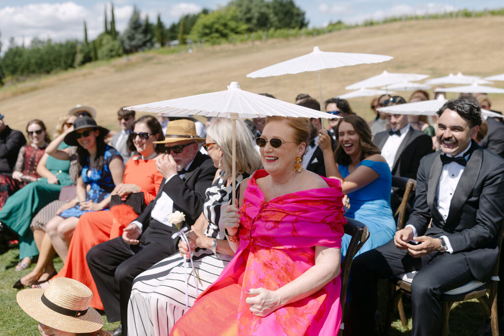A woman in a vibrant pink dress sits among other elegantly dressed guests holding white umbrellas at an outdoor wedding. The scene includes a grassy area and a scenic view of Lake Wanaka and mountains in the background under a partly cloudy sky.