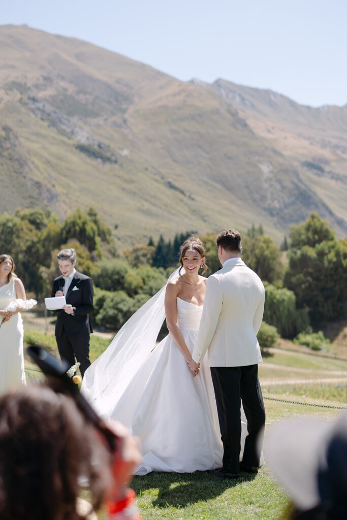 A friend of the bride and groom, in a black suit speaks into a microphone during an outdoor wedding ceremony at Rippon, Wanaka, with a bride and groom holding hands in white attire. Bridesmaids in white dresses with bouquets stand nearby, and the scenic backdrop includes vineyards, Lake Wanaka, and mountains under a clear sky.