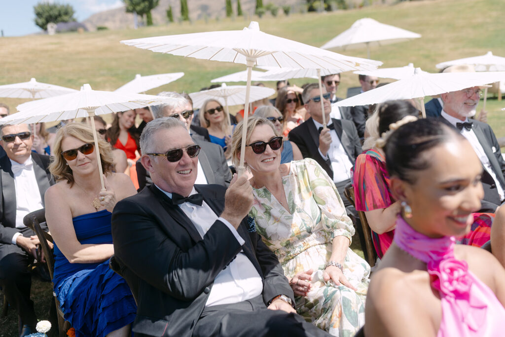 Guests seated on wooden chairs at an outdoor wedding, holding white umbrellas for shade. The attendees, dressed in elegant attire including suits and colourful dresses, smile and watch the ceremony. The backdrop features a grassy hill and distant mountains under a clear sky.