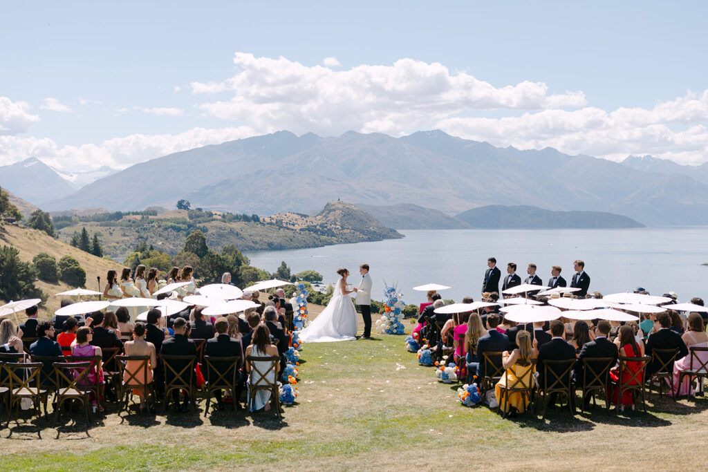 A wide view of an outdoor wedding ceremony at Rippon, Wanaka, with a bride and groom in white standing at the centre, surrounded by blue and orange floral arrangements. Guests seated on wooden chairs under white umbrellas watch, with Lake Wanaka and a mountain range visible in the background under a partly cloudy sky.