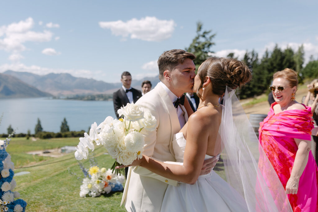 A bride and groom share a kiss during an outdoor wedding ceremony at Rippon, Wanaka. The bride, wearing a white strapless gown and veil, holds a bouquet of white roses and orchids, while the groom is dressed in a white suit with a black bow tie. They are surrounded by colourful floral arrangements, with Lake Wanaka and a mountain range visible in the background under a partly cloudy sky. Guests, including a woman in a vibrant pink dress, look on happily.