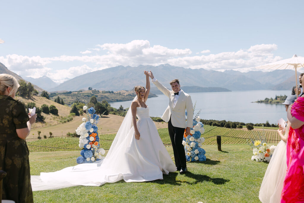 Bride and groom standing with their hands linked and arms raised, during their wedding ceremony outdoors at Rippon in Wanaka, New Zealand. The bride wears a strapless white gown with a long train and veil, while the groom wears a white jacket and black pants.  Behind them, vibrant blue, white, and orange floral arrangements frame the couple, with stunning views of Lake Wanaka, rolling hills, and distant mountains under a bright blue sky.