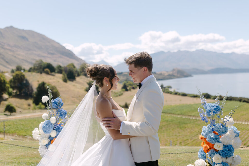 A bride and groom stand close together, smiling at each other during a wedding ceremony. The bride wears a white gown with a long veil, and the groom is in a white suit. They are surrounded by vibrant floral arrangements featuring blue and orange flowers, with a scenic backdrop of rolling hills, Lake Wanaka, and distant mountains under a partly cloudy sky.