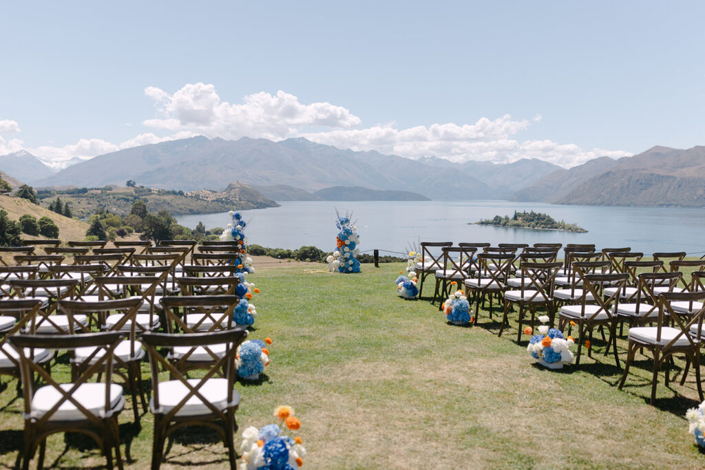 Outdoor wedding ceremony setup at Rippon, Wanaka, featuring rows of wooden cross-back chairs facing a picturesque lake and mountain backdrop. Vibrant floral arrangements in shades of blue, white, and orange line the aisle and create a colourful focal point at the altar. The lake and mountains stretch out under a clear blue sky with scattered white clouds.