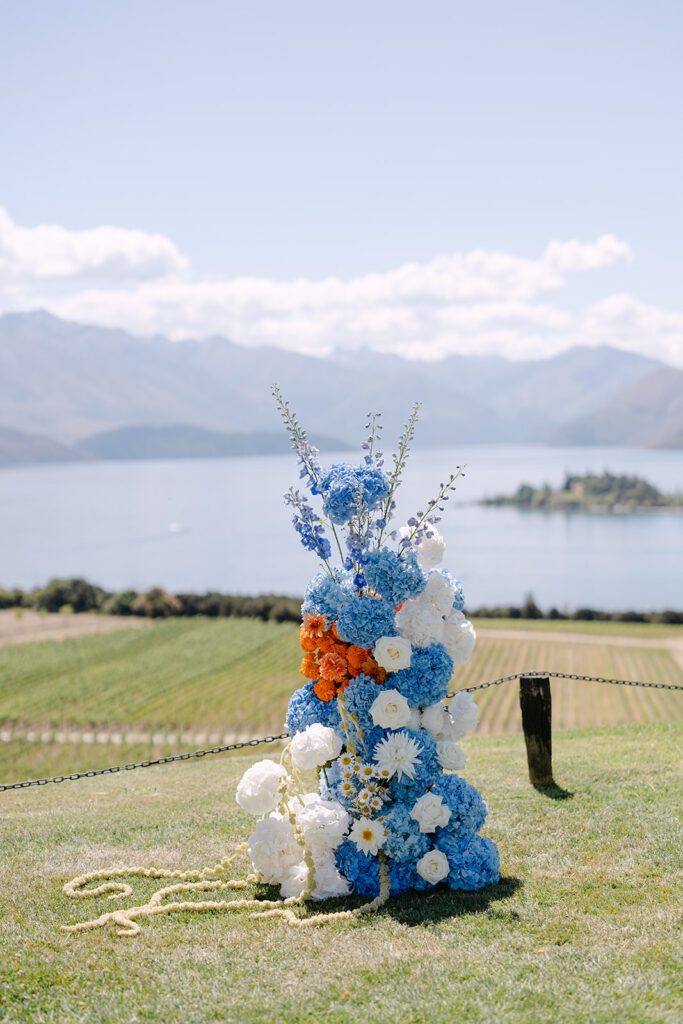 Vibrant, tall floral arrangement in shades of blue, white, and orange stand at the front of the aisle. An outdoor wedding setup at Rippon, Wanaka with the lake and mountains in the background.