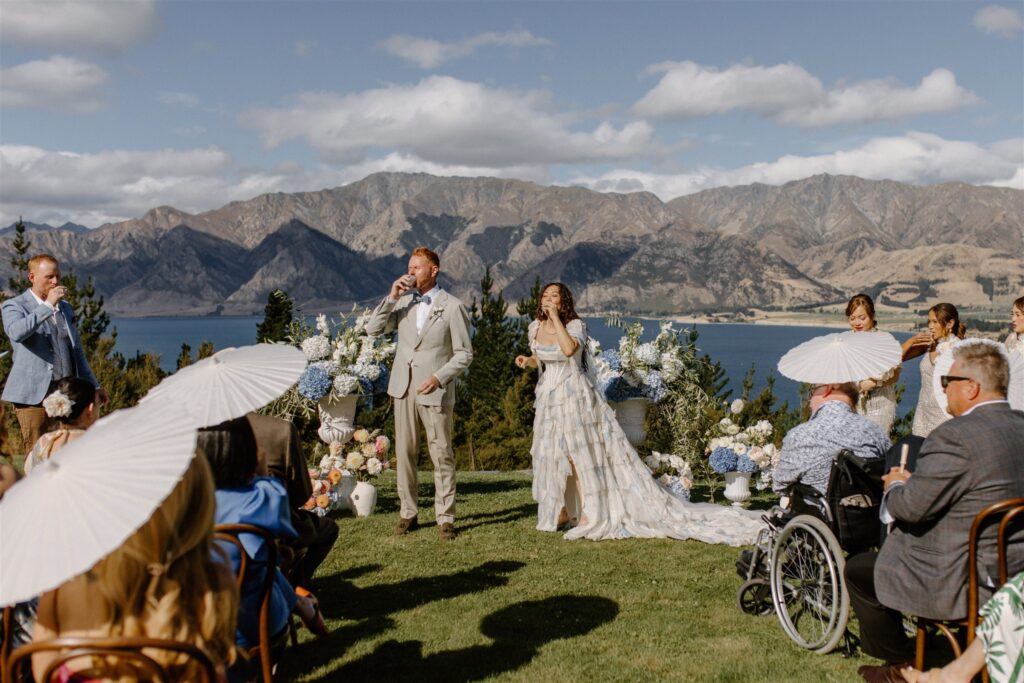 Bride and groom taking tequila shots during their outdoor wedding ceremony overlooking Lake Hawea with guests holding white parasols and mountain views in the background.