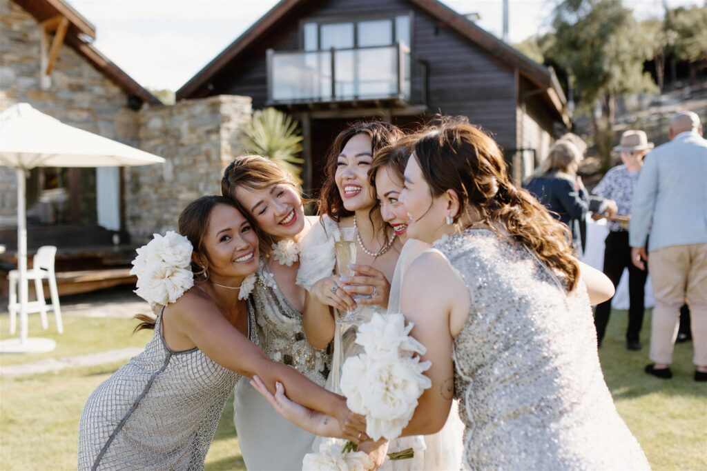 Five bridesmaids in sparkly dresses hugging and laughing together at an outdoor wedding reception with a rustic venue in the background.