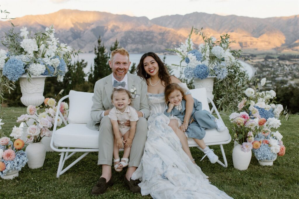 Smiling couple with two young children sitting on a white bench, surrounded by blue and pastel flowers with mountains and lake in the background.