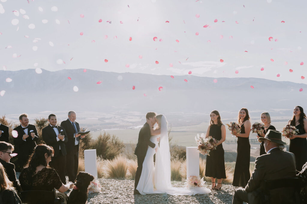 Bride and groom kissing during a mountaintop ceremony as pink petals float in the air, surrounded by smiling guests and a breathtaking mountain view.
