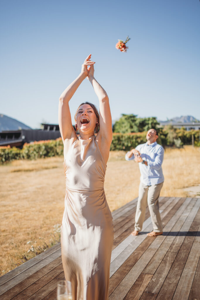 Guest laughing with arms raised as the wedding bouquet is tossed on a sunny Wanaka lawn.