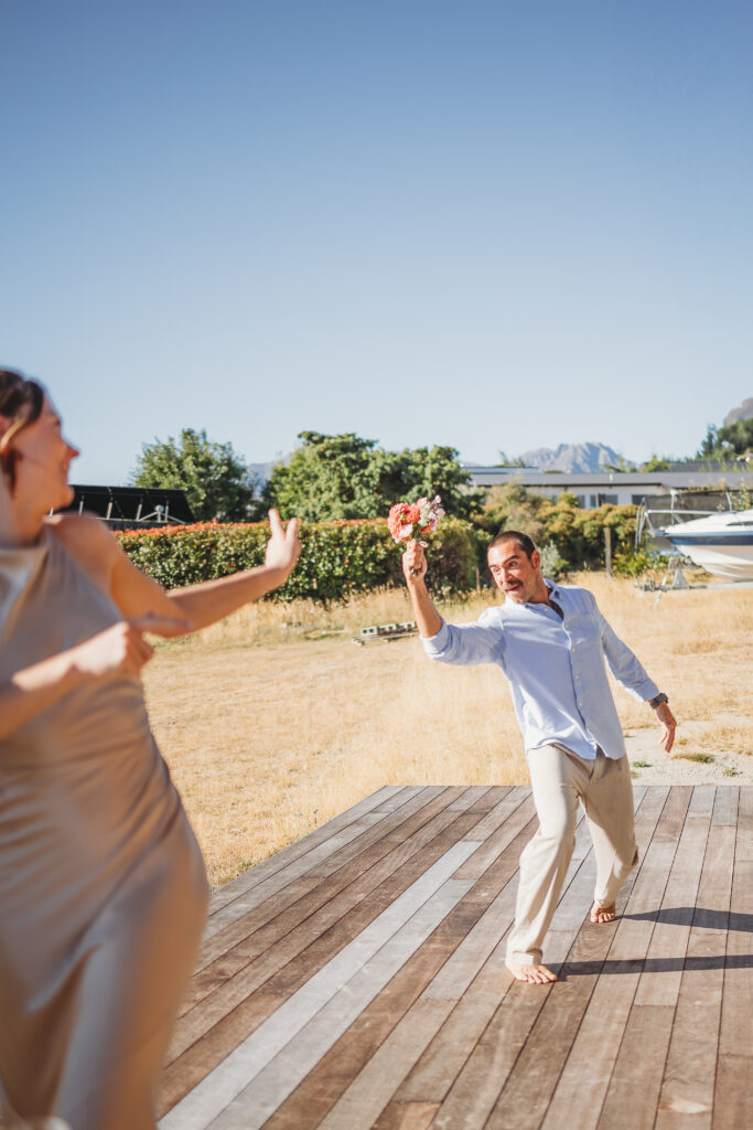 Man jokingly chasing a woman with a bouquet during a relaxed outdoor wedding celebration in Wanaka.
