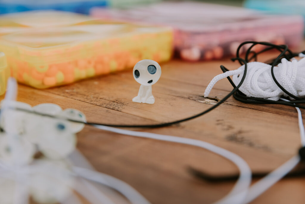 A tiny white figurine with big black eyes sits on a wooden table surrounded by colorful beads and crafting strings at a wedding activity station.