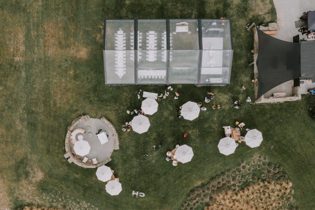 Drone shot from directly above the clear marquee and cocktail hour spaces, with guests mingling around parasols.