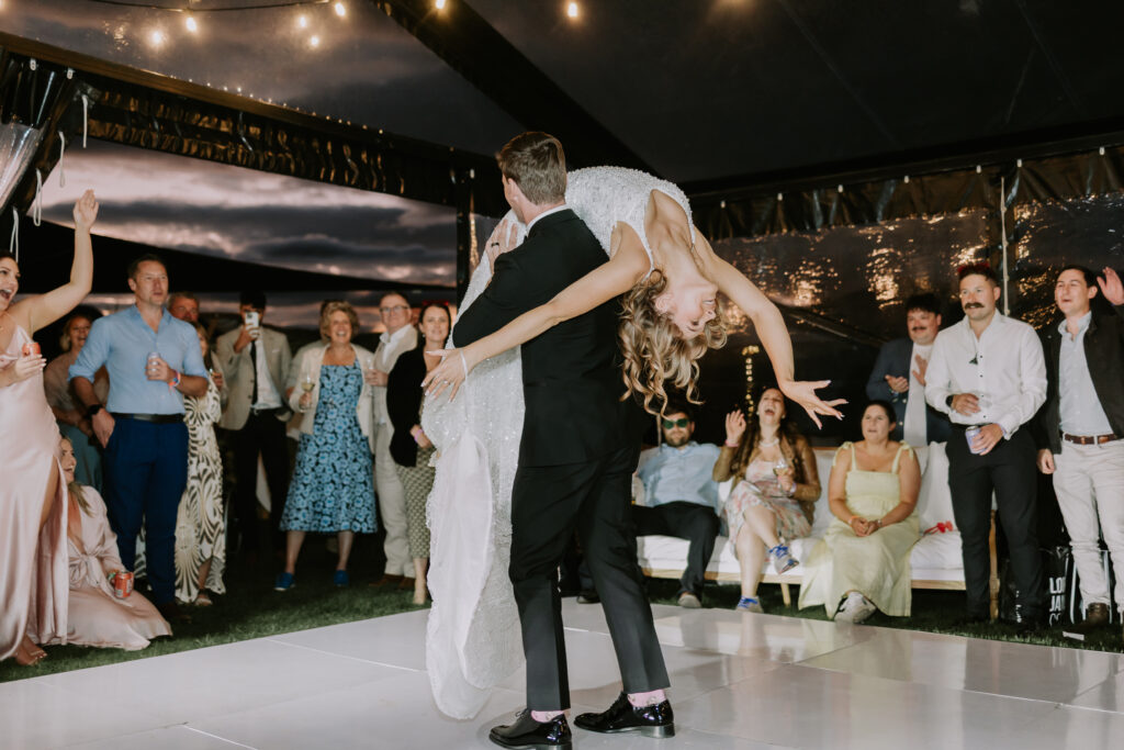 Bride and groom performing an energetic first dance surrounded by cheering guests under a marquee at sunset.