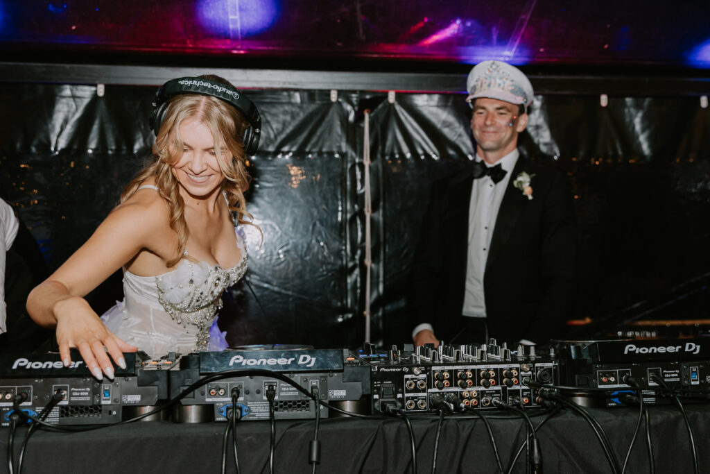 Bride DJing at the wedding afterparty with a big smile while the groom watches proudly in the rave-inspired marquee.