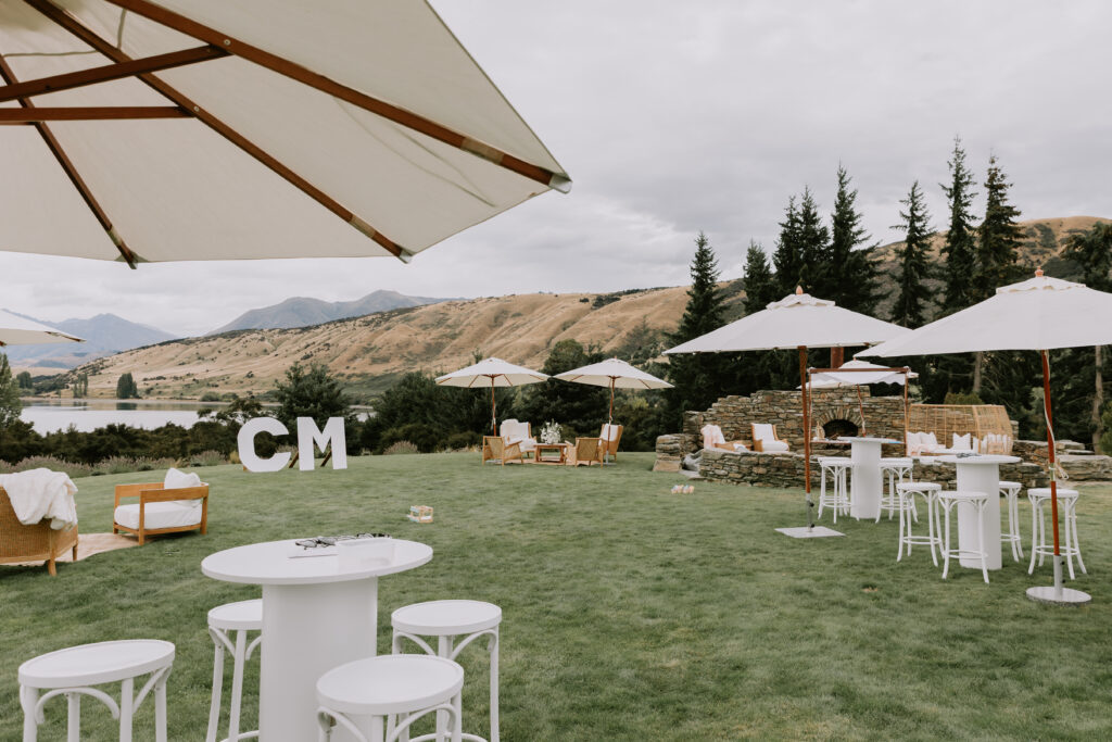 Outdoor cocktail lounge setup with white umbrellas, high tables, and large CM initials overlooking the lake and mountains.