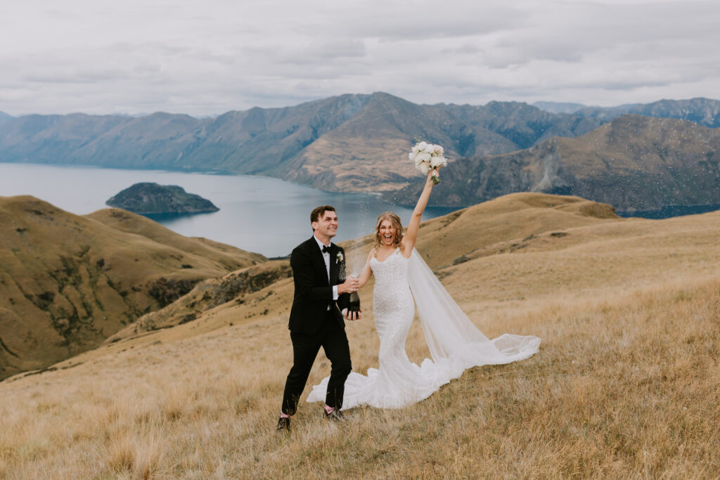 Bride and groom celebrate on a mountain top in Wanaka, New Zealand, popping champagne with stunning lake and alpine views in the background.