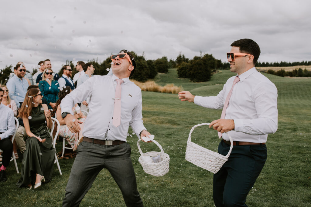Two groomsmen acting as fun “flower men,” throwing petals and laughing during the outdoor ceremony.