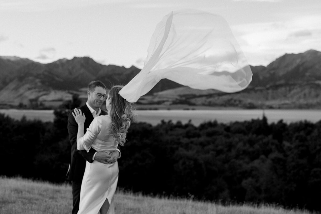 Bride’s veil blowing dramatically in the wind as couple embrace with Wanaka mountains in the background.