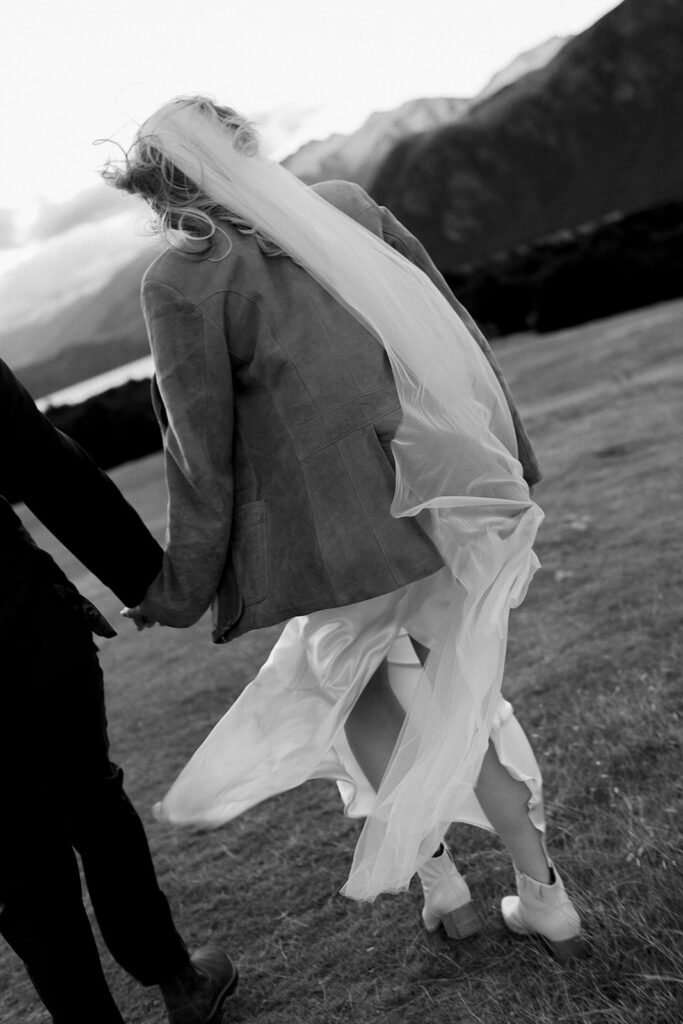 Close-up of bride’s veil flowing as the couple hold hands, walking across a windy alpine field.
