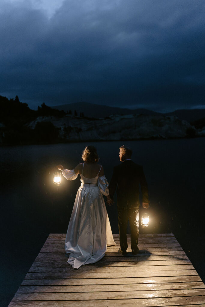 Bride and groom holding lanterns at night on a dock over Lake St Bathans, romantic moody elopement vibe.