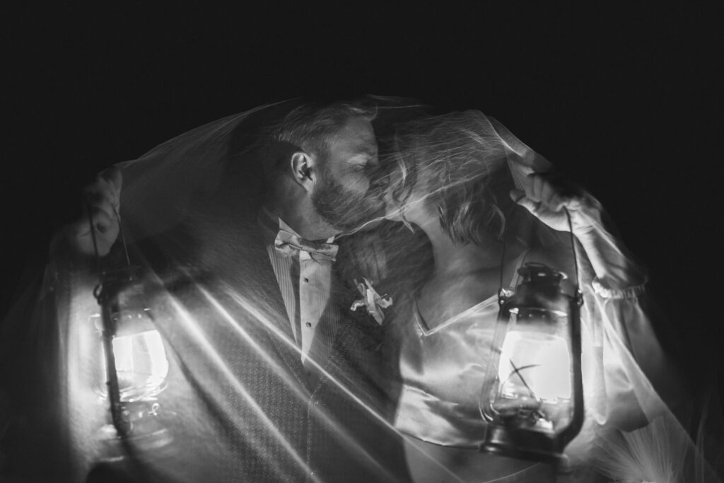 Black and white wedding photo of a bride and groom kissing under a veil while holding vintage lanterns glowing in the dark.