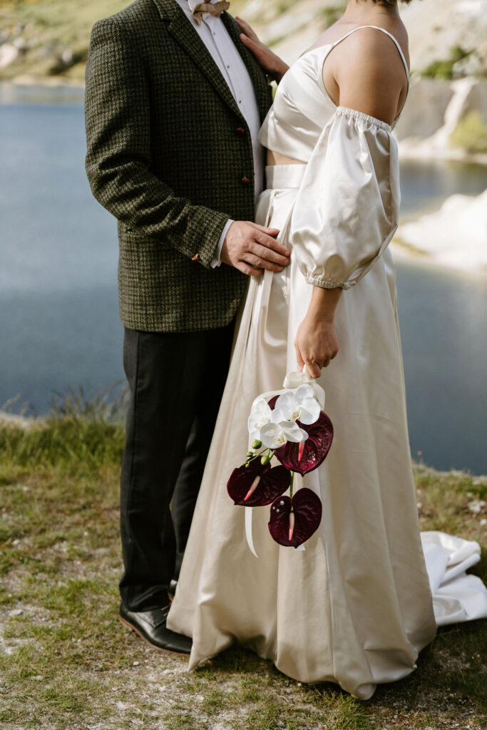 Close-up of bride’s silk gown and groom’s tweed jacket holding deep burgundy orchids at Lake St Bathans.