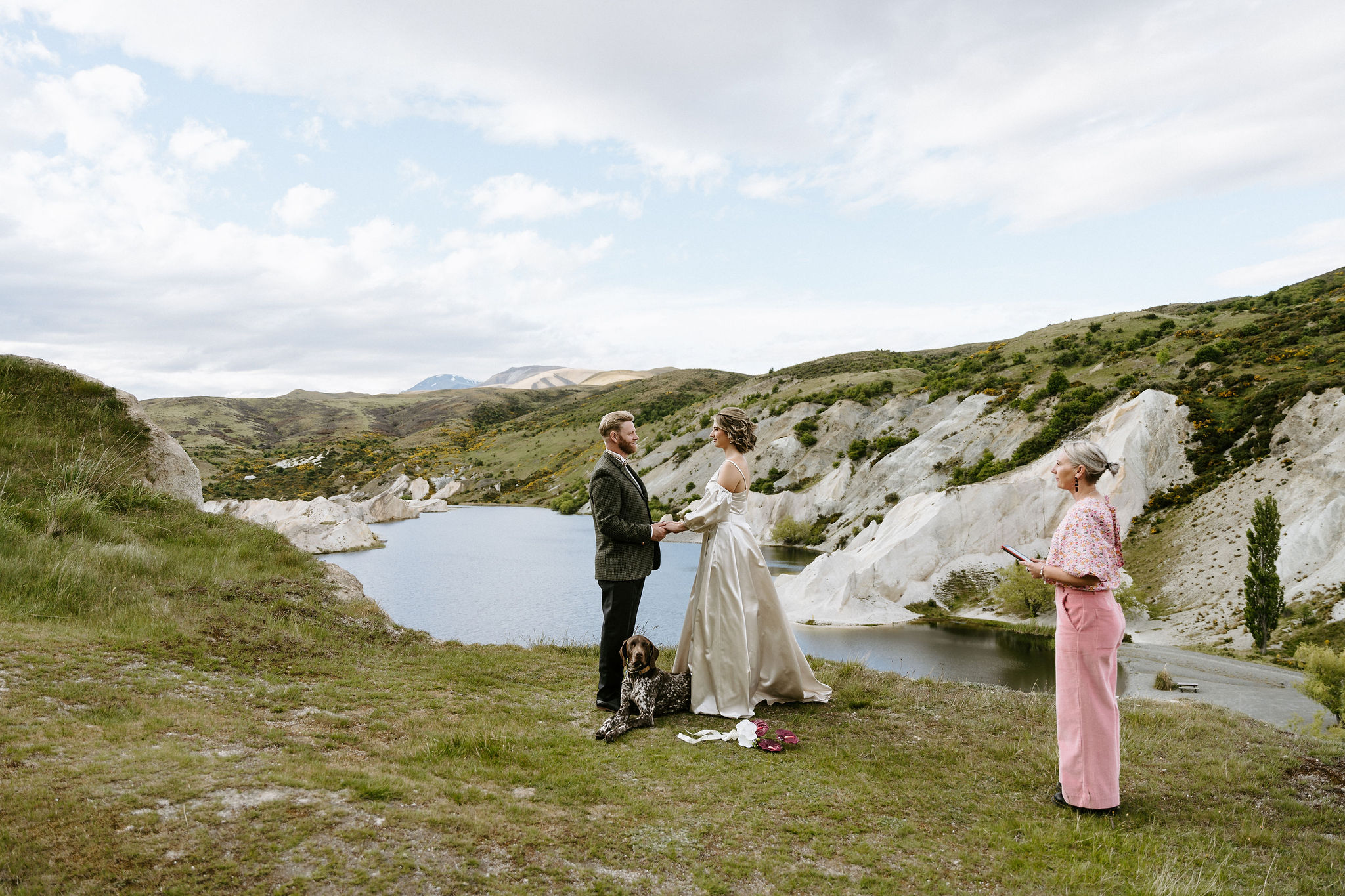 Bride and groom holding hands during an outdoor wedding ceremony overlooking a lake and rugged hills at St Bathans, New Zealand, with their dog lying at their feet and the celebrant standing nearby.