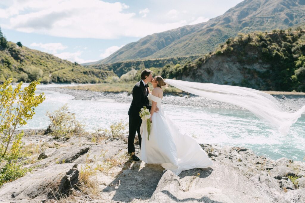 Bride and groom kissing by a river, veil blowing in the wind, Gibbston Valley