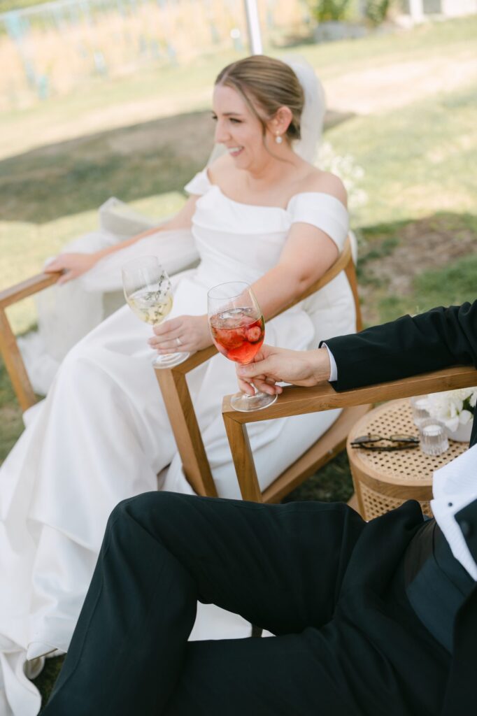 Bride laughing while seated with a drink, groom's hand reaching in beside her