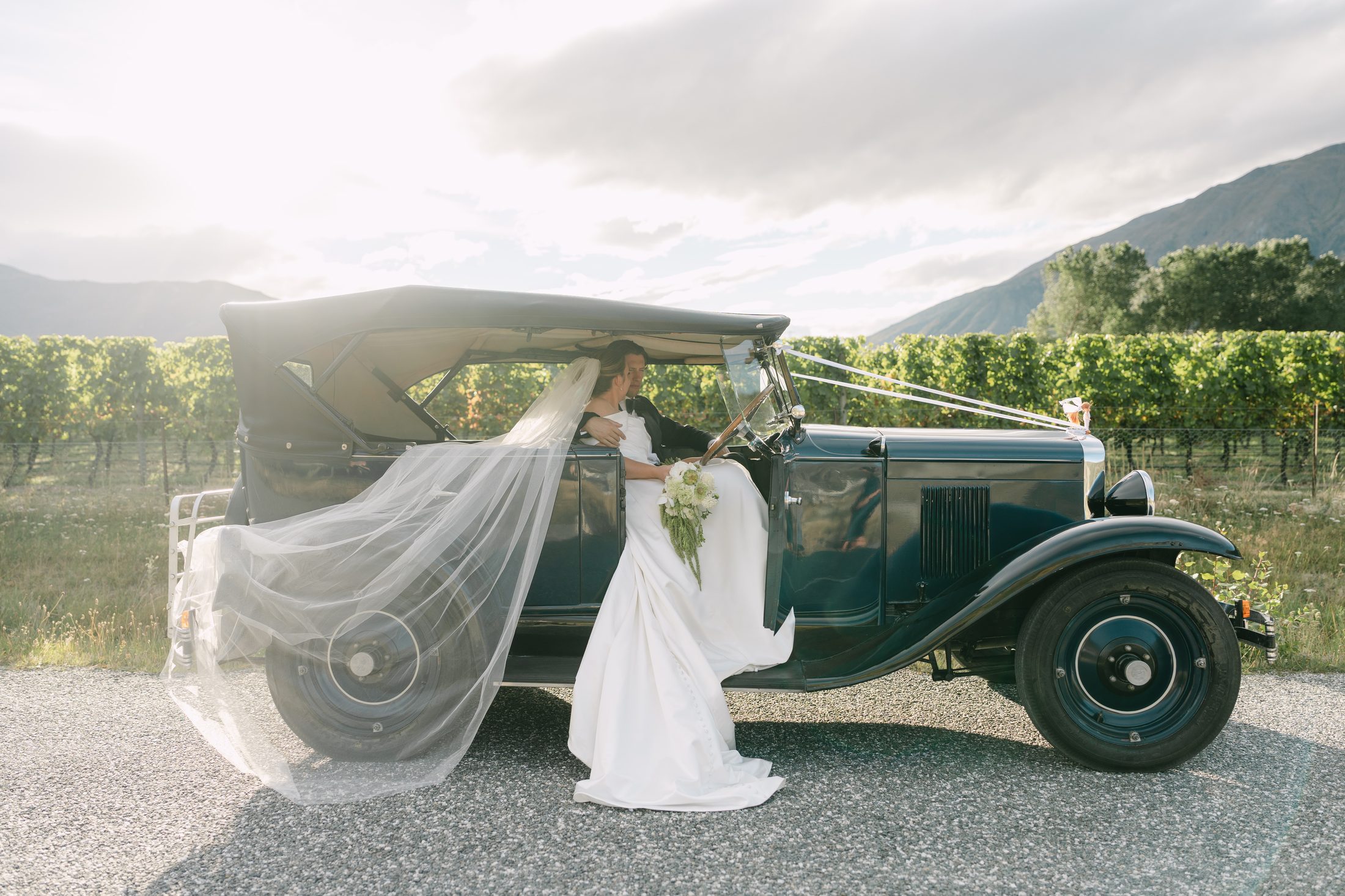 Bride and groom seated in a vintage car with veil flowing, Gibbston vineyard behind