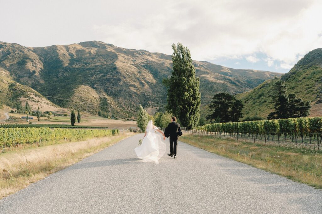Bride and groom walking away hand in hand down a vineyard road, Central Otago mountains behind